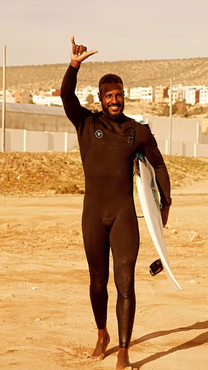 Momo holding his surfboard on the beach, smiling