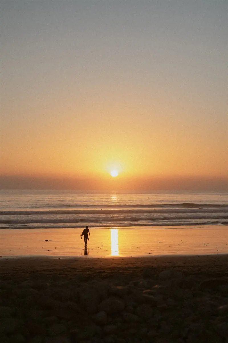 Surfer walking along the beach at sunset with a board under one arm